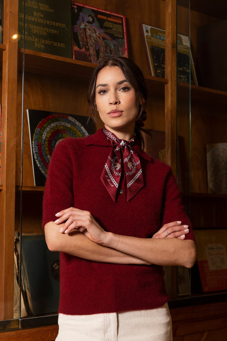 Smiling woman wearing a brown long-sleeve polo shirt and a pink wrist scarf, holding a red apple in a bright, modern interior.
